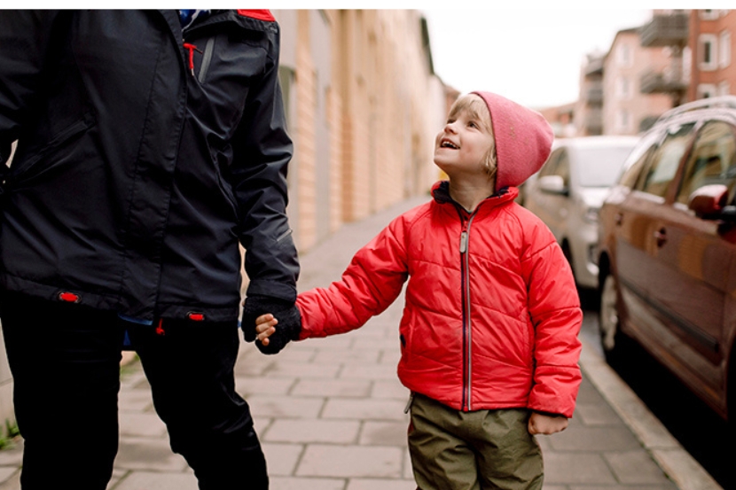 Alakoukuikäinen punaiseen takkiin ja pipoon pukeutunut lapsi kulkee kadulla pitäen kiinni aikuisen kädestä ja katsoen aikuiseen.