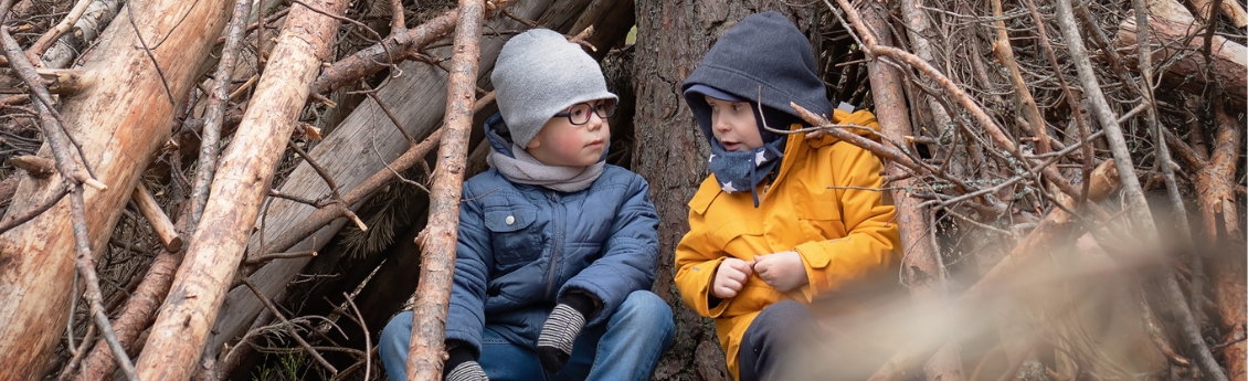Kaksi välikausivaatteisiin pukeutunutta leikki-ikäistä lasta istuu vierekkäin puumajassa puunrunkoon nojaten.