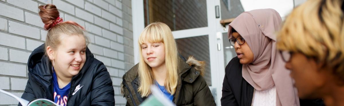 Neljä nuorta istuu rinnakkain ulkona vaalean tiilirakennuksen edustalla ja katsoo toisiinsa. Yhden nuoren kädessä on koulukijalta näyttävä kirja.